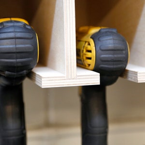May include: Close-up of three yellow and black cordless drills stored in a wooden rack. The drills are held in place by the rack's design, which allows for easy access and organization.