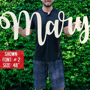 May include: A person holds a large, white wooden sign with the name "Mary" written in a cursive font. The sign is 48 inches tall. The person is standing in front of a green hedge.