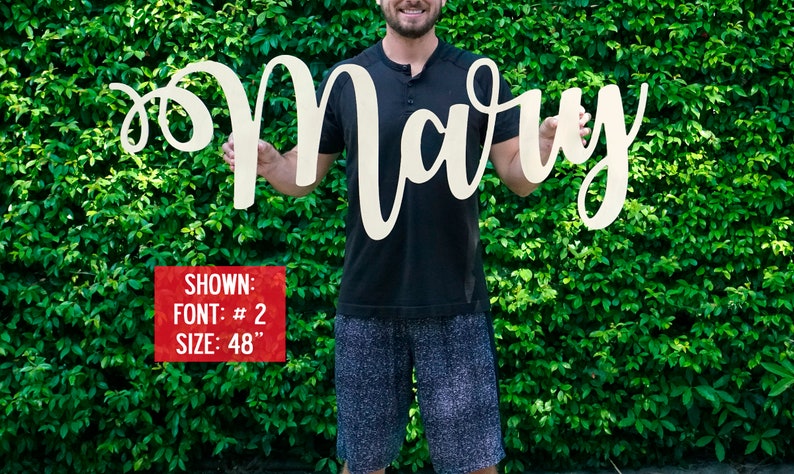 May include: A person holds a large, white wooden sign with the name "Mary" written in a cursive font. The sign is 48 inches tall. The person is standing in front of a green hedge.