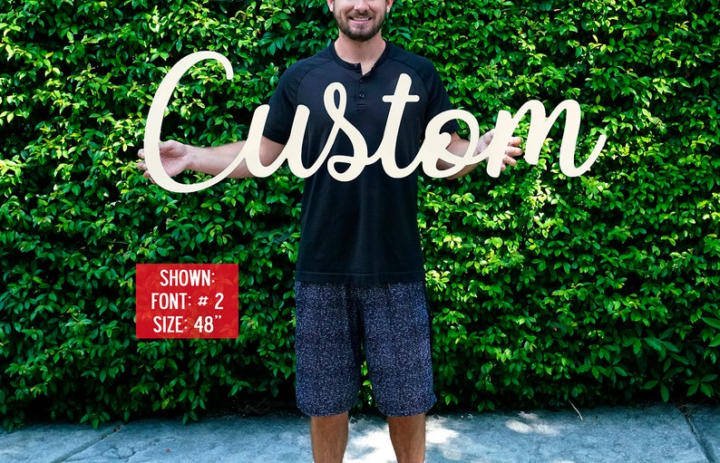 May include: A person holds a white wooden sign that says "Custom" in a cursive font. The sign is 48 inches long. Shown: Font #2, Size: 48"