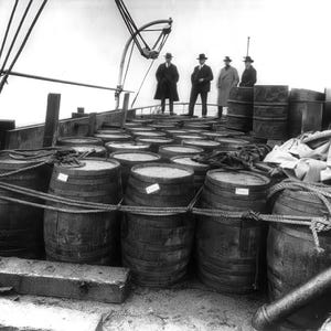 May include: A black and white photo of a ship's deck with numerous wooden barrels stacked and secured with rope.  Four men in suits stand on the deck, looking out at the water.