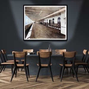 Op de afbeelding: Een ingelijste zwart-witfoto van een scheepsdek met houten banken en uitzicht op de oceaan. De foto hangt aan een donkergrijze muur boven een eettafel met zes zwarte stoelen.