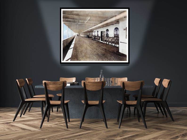 Op de afbeelding: Een ingelijste zwart-witfoto van een scheepsdek met houten banken en uitzicht op de oceaan. De foto hangt aan een donkergrijze muur boven een eettafel met zes zwarte stoelen.