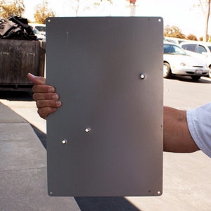 May include: A gray metal sign with four holes drilled into it. The sign is being held by a person's hand.