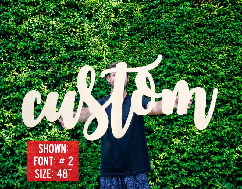 May include: A person holds up a large, white wooden sign that says "custom" in a cursive font. The sign is 48 inches tall. Shown: Font # 2, Size: 48"