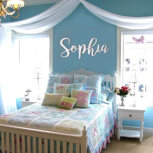 May include: A white wooden sign with the name "Sophia" in cursive script, mounted on a light blue wall above a white bed with a blue and white floral patterned quilt and pillows. The room is decorated with a white canopy and a white nightstand with a lamp.