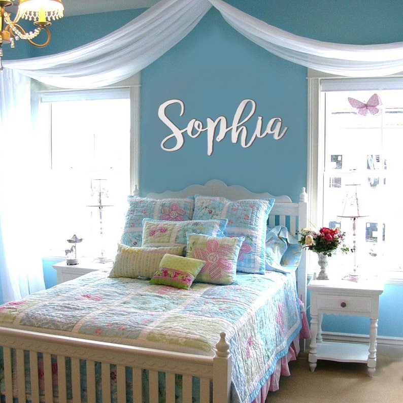 May include: A white wooden sign with the name "Sophia" in cursive script, mounted on a light blue wall above a white bed with a blue and white floral patterned quilt and pillows. The room is decorated with a white canopy and a white nightstand with a lamp.