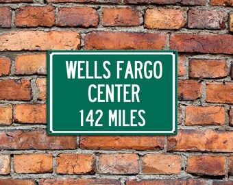 Puede incluir: Un letrero verde con texto blanco que dice "Wells Fargo Center 142 Miles" montado en una pared de ladrillo.