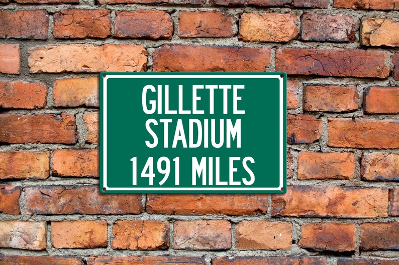 May include: A green sign with white text that reads "Gillette Stadium 1491 Miles". The sign is mounted on a brick wall.