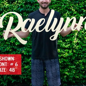 May include: A white wooden sign with the name "Raelynn" written in a cursive font. The sign is 48 inches tall and is being held by a person. The sign is shown in font #6.
