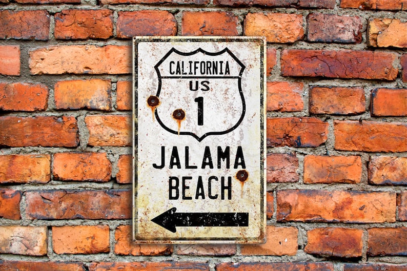 May include: A white metal sign with black text and a black arrow pointing left. The sign reads "California US 1 Jalama Beach".