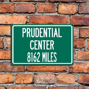 May include: A green sign with white text that reads "Prudential Center 8162 Miles" mounted on a brick wall.