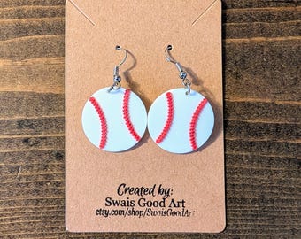 May include: A pair of baseball-themed earrings. Each earring is a white circle with red stitching, resembling a baseball. The earrings hang from silver-colored hooks. The earrings are displayed on a brown card with text that reads "Created by: Swais Good Art etsy.com/shop/SwaisGoodArt:"