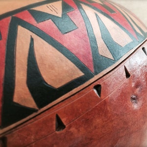 May include: A brown gourd with a geometric design painted in red, black, and orange. The gourd has a smooth surface and is decorated with a band of geometric shapes around the top.