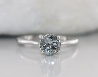 May include: A silver ring with a round, light blue gemstone set in a four-prong setting.