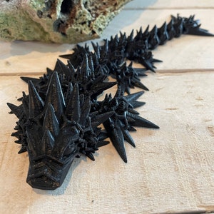 May include: A black, 3D printed, spiky creature with a long tail. The creature has a sharp, pointed head and a textured, scaly body.