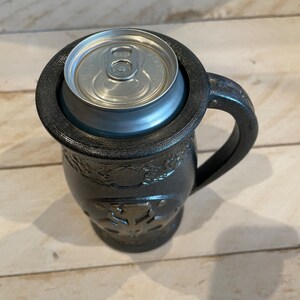May include: A black ceramic mug with a handle and a decorative design. A silver can is sitting inside the mug.