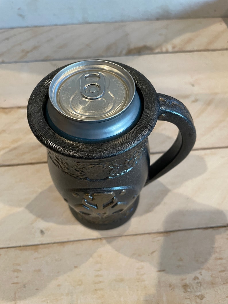 May include: A black ceramic mug with a handle and a decorative design. A silver can is sitting inside the mug.