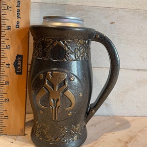 May include: A black and gold ceramic mug with a Mandalorian symbol on the front. The mug has a handle and a textured surface.