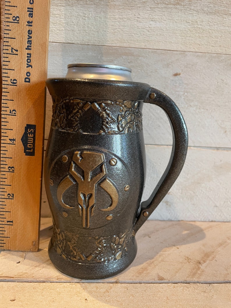 May include: A black and gold ceramic mug with a Mandalorian symbol on the front. The mug has a handle and a textured surface.