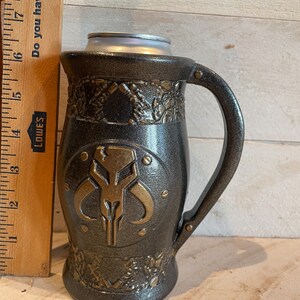 May include: A black and gold ceramic mug with a Mandalorian symbol on the front. The mug has a handle and a textured surface.