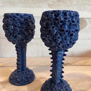 May include: Two dark blue goblets with a skull-shaped design. The goblets are made of a textured material and have a unique, skeletal stem.