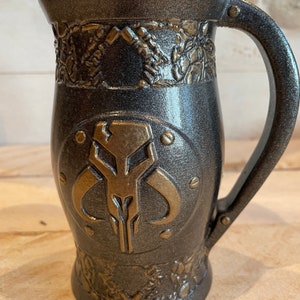 May include: A dark gray ceramic mug with a gold Mandalorian symbol and decorative details. The mug has a handle and a rounded bottom.