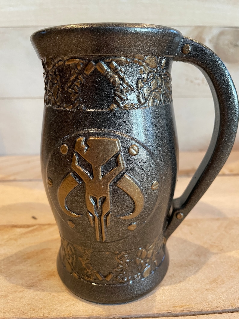 May include: A dark gray ceramic mug with a gold Mandalorian symbol and decorative details. The mug has a handle and a rounded bottom.
