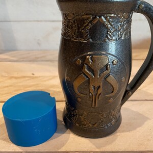 May include: A black and gold ceramic mug with a Mandalorian symbol on the front. The mug has a handle and a textured design.