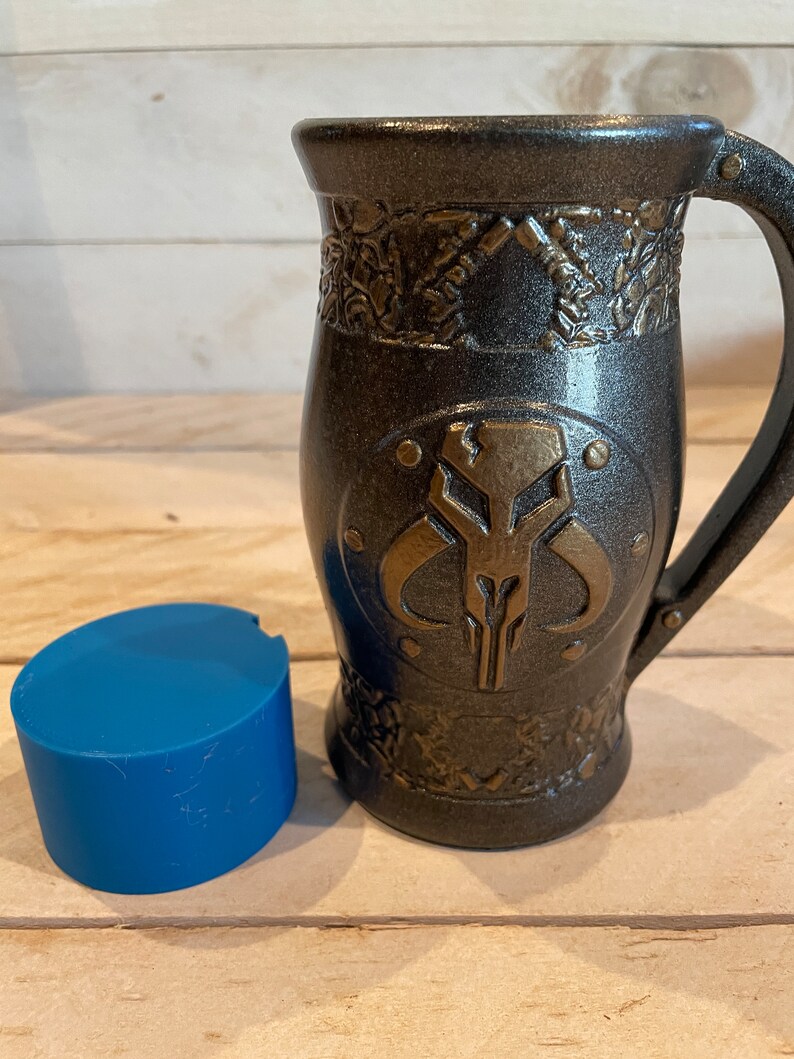 May include: A black and gold ceramic mug with a Mandalorian symbol on the front. The mug has a handle and a textured design.
