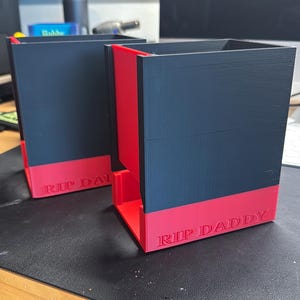Puede incluir: Dos contenedores rectangulares negros y rojos. Los contenedores tienen un cuerpo negro con una base y un borde rojos. El texto "RIP DADDY" está impreso en blanco en la base roja de cada contenedor. Los contenedores están sobre una superficie negra.