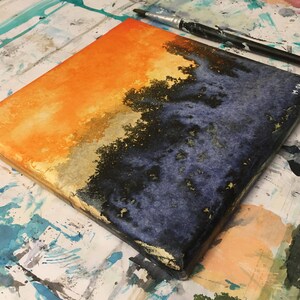 May include: A square canvas painting with a horizontal division of orange and blue. The top half is a bright orange with a gradient to a lighter yellow. The bottom half is a dark blue with black and gold accents. The canvas is sitting on a white surface with paint splatters.