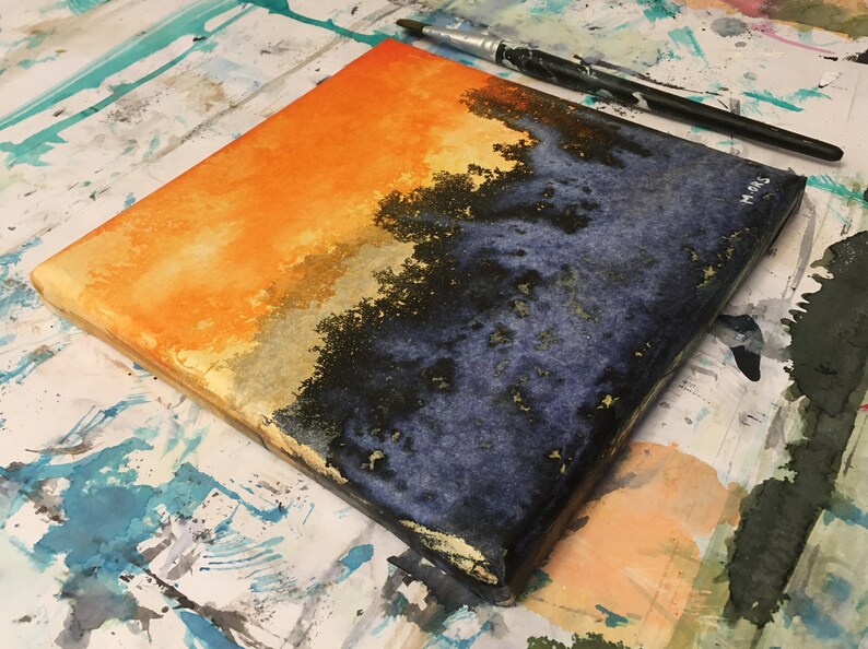 May include: A square canvas painting with a horizontal division of orange and blue. The top half is a bright orange with a gradient to a lighter yellow. The bottom half is a dark blue with black and gold accents. The canvas is sitting on a white surface with paint splatters.