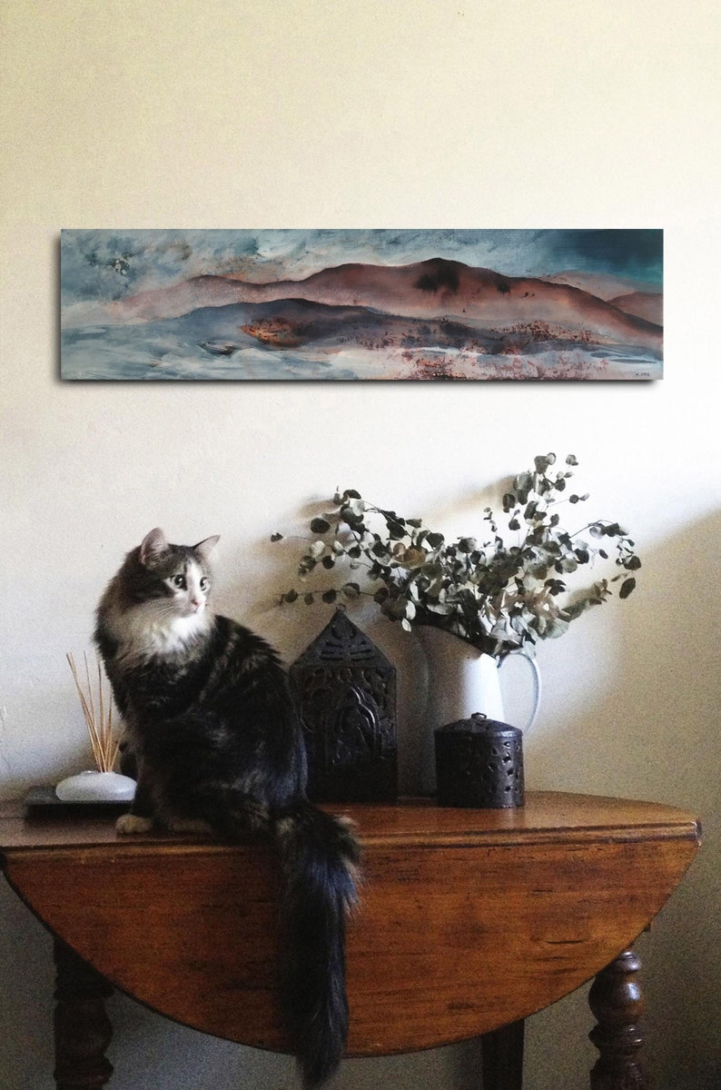 May include: A brown wooden table with a cat sitting on it. The table has a rounded top and turned legs. There is a vase of dried eucalyptus branches, a black lantern, and a black candle holder on the table. A painting of a landscape with blue and brown hues hangs on the wall behind the table.