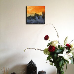 May include: A small abstract painting with a blue and orange color scheme. The painting is hanging on a white wall above a table with a vase of flowers and decorative objects.