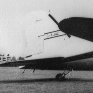 May include: Black and white image of an airplane's tail and wing. The tail features the text "G-A.JHZ". The wing is dark, and the plane appears to be on a grassy surface. The image is in black and white.