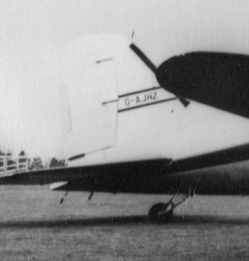 May include: Black and white image of an airplane's tail and wing. The tail features the text "G-A.JHZ". The wing is dark, and the plane appears to be on a grassy surface. The image is in black and white.
