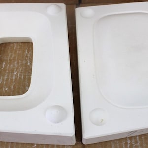 May include: Two white ceramic molds, one with a rectangular opening and the other solid. Each mold has a rounded shape and small circular indentations. The molds are likely for casting or forming objects.