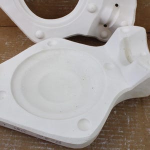 May include: Two white, irregularly shaped ceramic molds. One has a large circular opening, and the other has a circular indentation. Both have rounded protrusions and sit on a brown cardboard surface.
