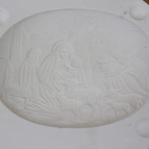 May include: A white, oval-shaped ceramic mold featuring a relief scene of the Nativity. The scene includes figures of Mary, Joseph, and the baby Jesus, along with sheep and the three wise men. The mold has rounded corners and three raised circular details.