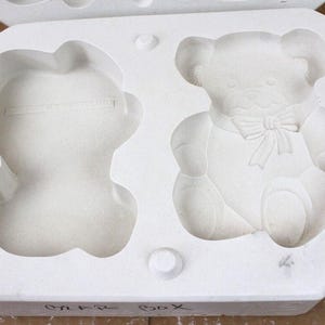 May include: A white plaster mold with two teddy bear designs. One bear is a full figure with a bow tie, the other is a partial figure. The mold is labeled "CAKE BOX" in black marker.