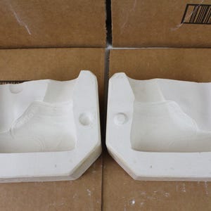 May include: Two white ceramic molds shaped like sneakers. Each mold has a detailed outline of a shoe, including the laces and sole. The molds are set against a neutral background.