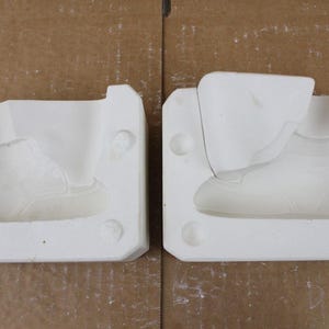 May include: Two white ceramic molds, each shaped like a high-top sneaker, are displayed side-by-side. The molds are detailed with the shoe's design, including the sole, laces, and ankle support. The background is a light brown surface.