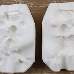 May include: Two white ceramic molds, each featuring a seated angel with detailed wings. The molds are oval-shaped with additional decorative elements. Ideal for crafting or decorative projects.