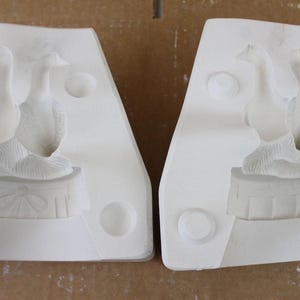 May include: Two-part white ceramic mold featuring a pair of geese. The geese are perched on a decorative base with a textured surface. The mold is designed for creating ceramic figurines or decorative items.