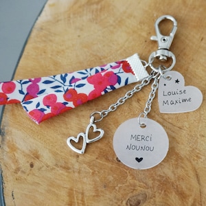Peut inclure: Un porte-clés avec un mousqueton argenté, une bande de tissu à motif floral rouge et bleu, deux breloques cœurs argentés et deux disques argentés. Un disque porte l'inscription "MERCI NOUNOU" avec un cœur, l'autre "Louise Maxime".