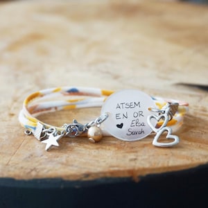 Peut inclure: Un bracelet avec un bracelet à motifs blancs, jaunes et bleus. Il comporte un charm en argent avec le texte "ATSEM EN OR Elsa Sarah" et des symboles de cœur, ainsi que des breloques en forme d'étoile et de cœur.