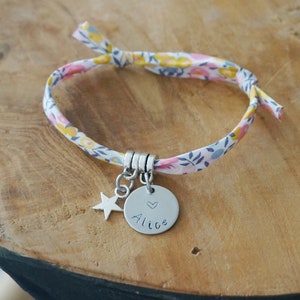 Peut inclure: Un bracelet en tissu blanc avec un motif floral rose, jaune et bleu. Le bracelet a un charme argenté avec le nom "Alice" et un petit cœur.