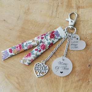 Peut inclure: Un porte-clés argenté avec un ruban en tissu floral, un charme en forme de cœur argenté avec un motif d'arbre, un charme rond argenté avec le texte "Maman de trois", et un charme en forme de cœur transparent avec le texte "Lukas Maya Oliver".