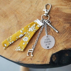 Peut inclure: Un porte-clés composé d'une bande de tissu jaune moutarde à motif floral blanc, d'une breloque flûte en métal argenté, d'une note de musique et d'un disque translucide gravé du texte Léa Flûtiste en Or avec un petit cœur.